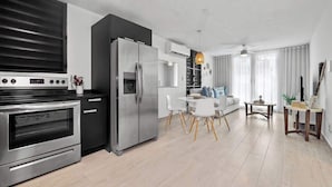 Une cuisine moderne dotée d’un four et d’un réfrigérateur encastrés, un coin repas avec une table et des chaises blanches, et un espace de vie comprenant un canapé et une table basse.