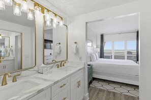 Habitación de hotel con un espejo grande, dos lavamanos, una cama y vista al mar.