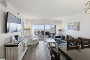 Una sala de estar moderna con sofá, mesa de centro y televisor, un comedor con sillas y vistas amplias a través de grandes ventanas.