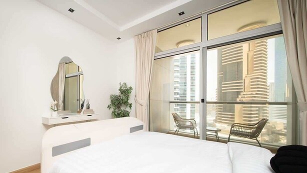 A modern hotel room with a large bed, a sitting area with a chair, a large window with a view of tall buildings, and a mirror on the wall. - Dubai Marina Apt w/ Pool, Gym & Balcony Views (Dubai)