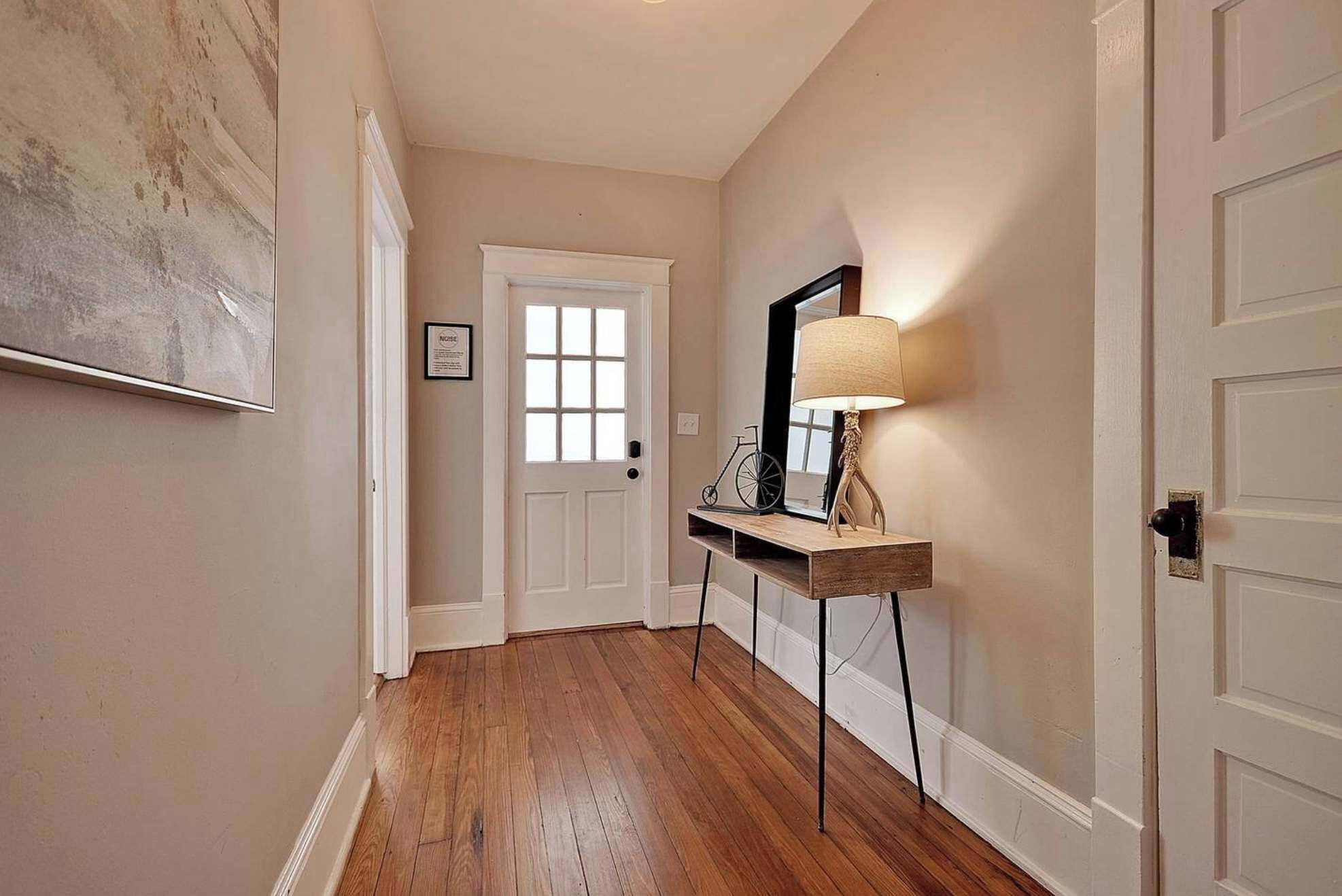 A hallway with wooden flooring, a white door, a framed picture on the wall, and a lamp on a wooden console table.