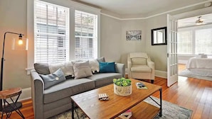 A living room with a grey sofa, a wooden coffee table, and a white armchair. There are large windows with blinds, a floor lamp, and a ceiling fan. - Spacious 3BR Home, Walk to King St! (Charleston)