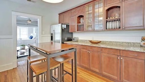 A kitchen with wooden cabinets, a stainless steel refrigerator, a granite countertop, and a dining area with a wooden table and chairs. - Spacious 3BR Home, Walk to King St! (Charleston)