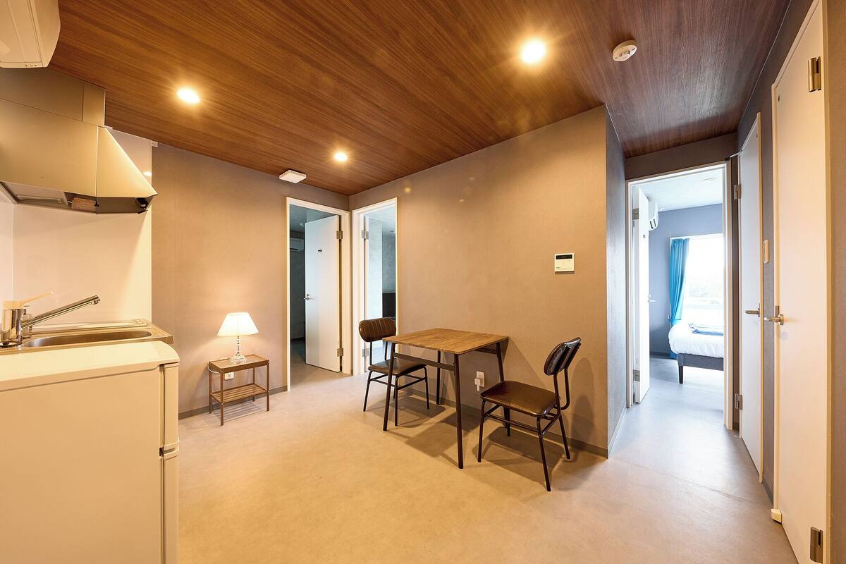 Et kompakt køkken med vask, et lille bord med stole og en lampe. Der er trælofter og en dør, der fører til et andet rum.
