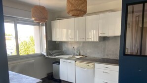 A modern kitchen with white cabinets, a built-in dishwasher, and a window with a view of greenery outside. - Family housing - Lyon 6e/Villeurbanne - Charpennes (Villeurbanne)