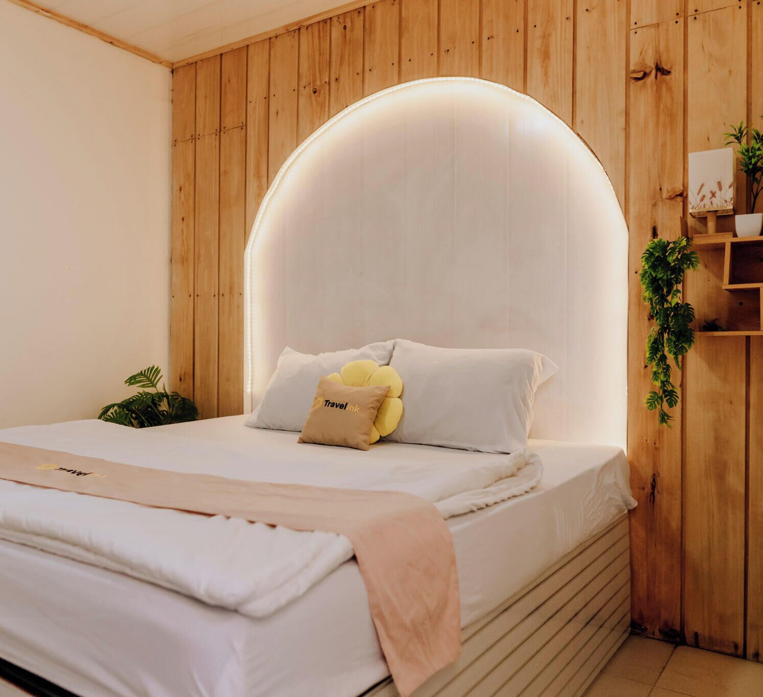 A bedroom with a wooden headboard, a bed with white bedding, a bedside plant, and a shelf with decorative items.