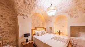 A bedroom with a stone wall, a bed with a wooden headboard, a bedside table with a lamp, and a small shelf with towels. - Old Town Şanlıurfa (Şanlıurfa)
