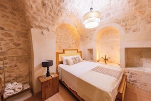 Quarto com parede de pedra, cama com cabeceira de madeira, mesa de cabeceira com luminária e prateleira pequena com toalhas.