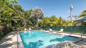Una piscina rectangular con borde de azulejos, rodeada de sillas de descanso y una valla verde.