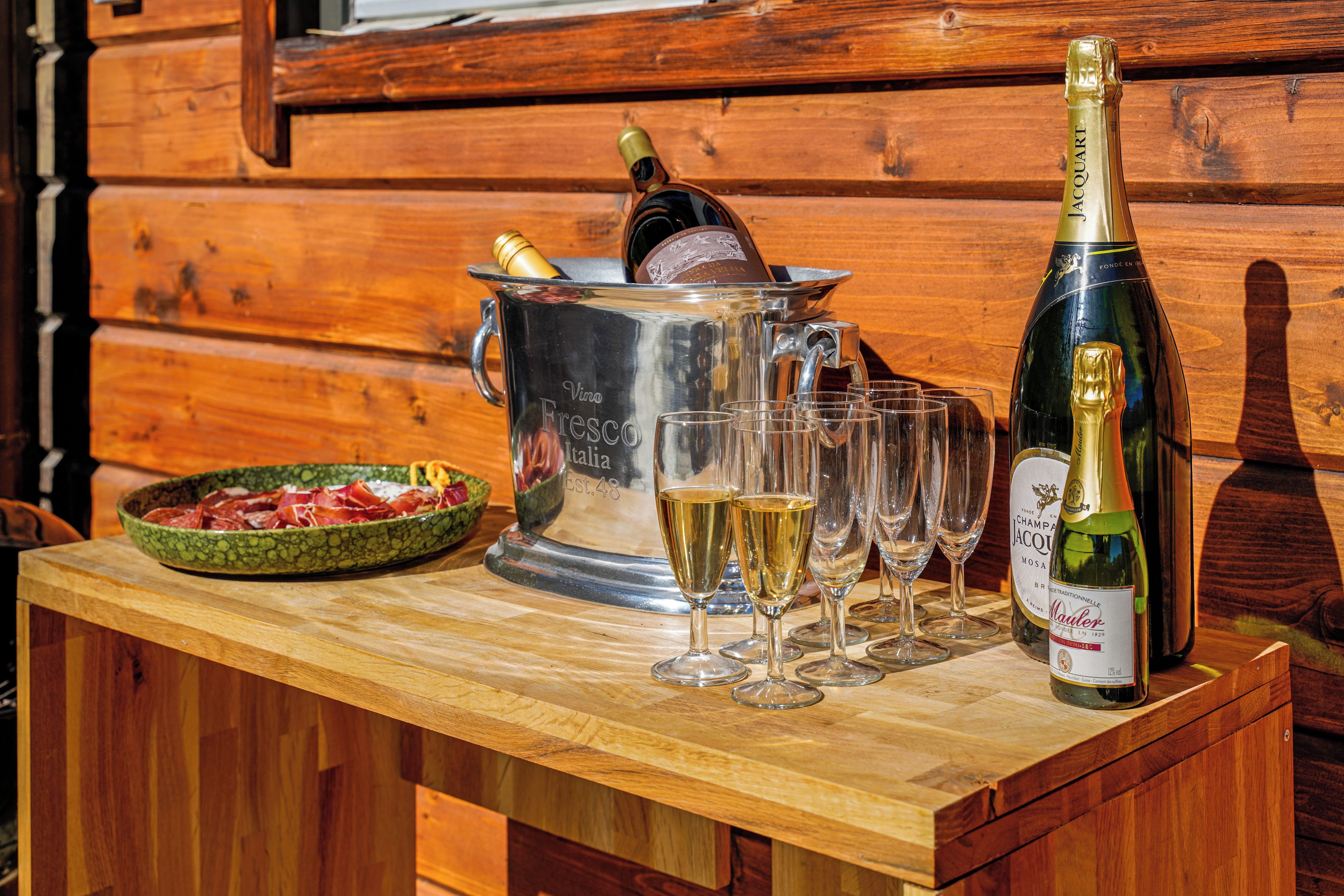 A wooden table with a wine bucket, glasses, a bottle of champagne, and a bowl of cured meats.