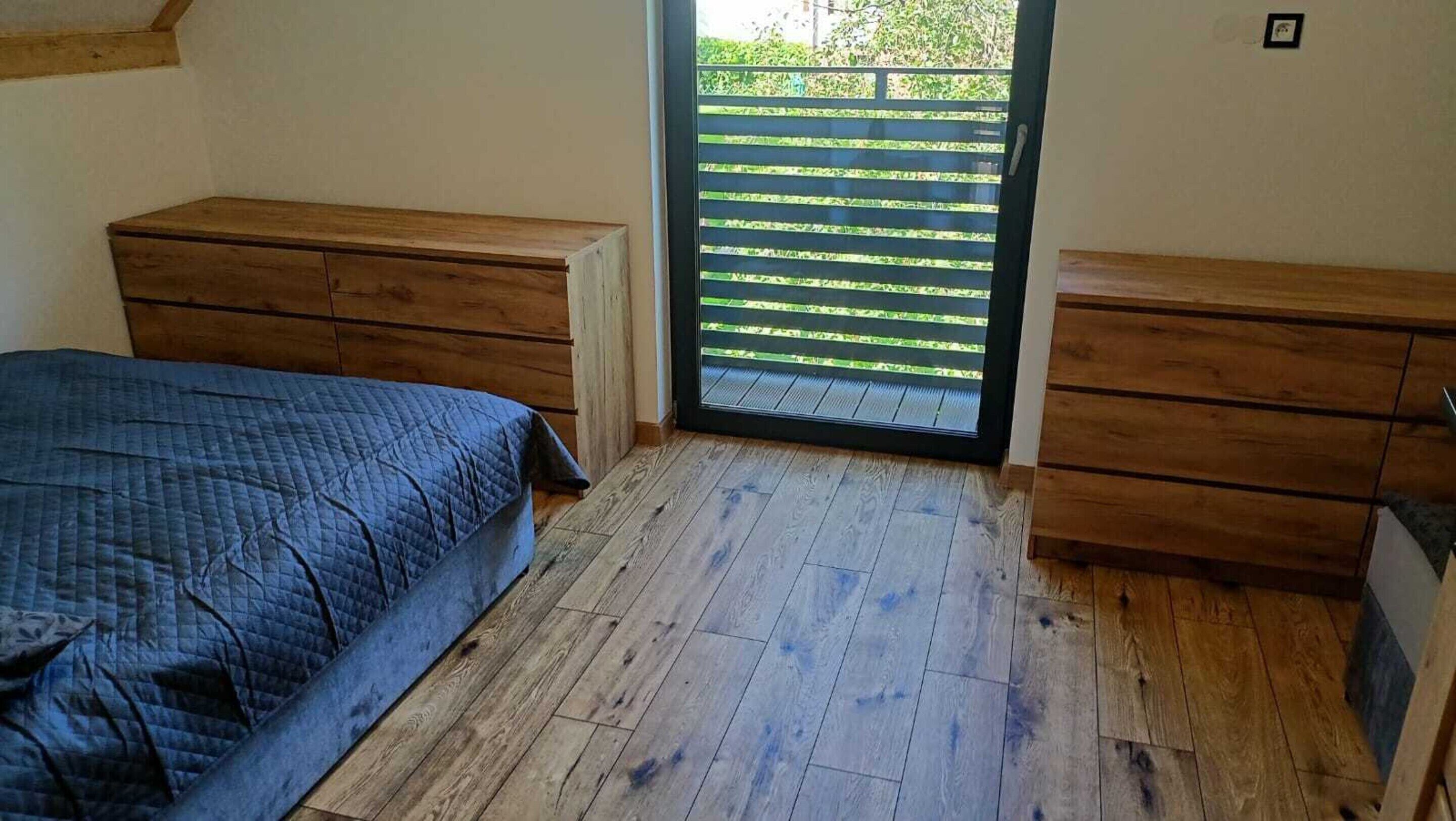 A bedroom with a bed, two wooden dressers, a window with blinds, and a wooden floor.