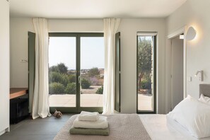A bedroom with a bed, a nightstand, a window with curtains, and a view of the outdoors.