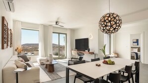 A modern living room with a sofa, dining table, and chairs. There is a TV mounted on the wall, a ceiling fan, and a large window with a view of the outdoors. - Ampelas 4BR Villa, Private Pool, To Ktima Paros (Paros)