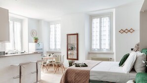 A modern bedroom with a bed, green upholstered headboard, a small desk with a chair, and a window with sheer curtains. - Dinghy Studio (Como)