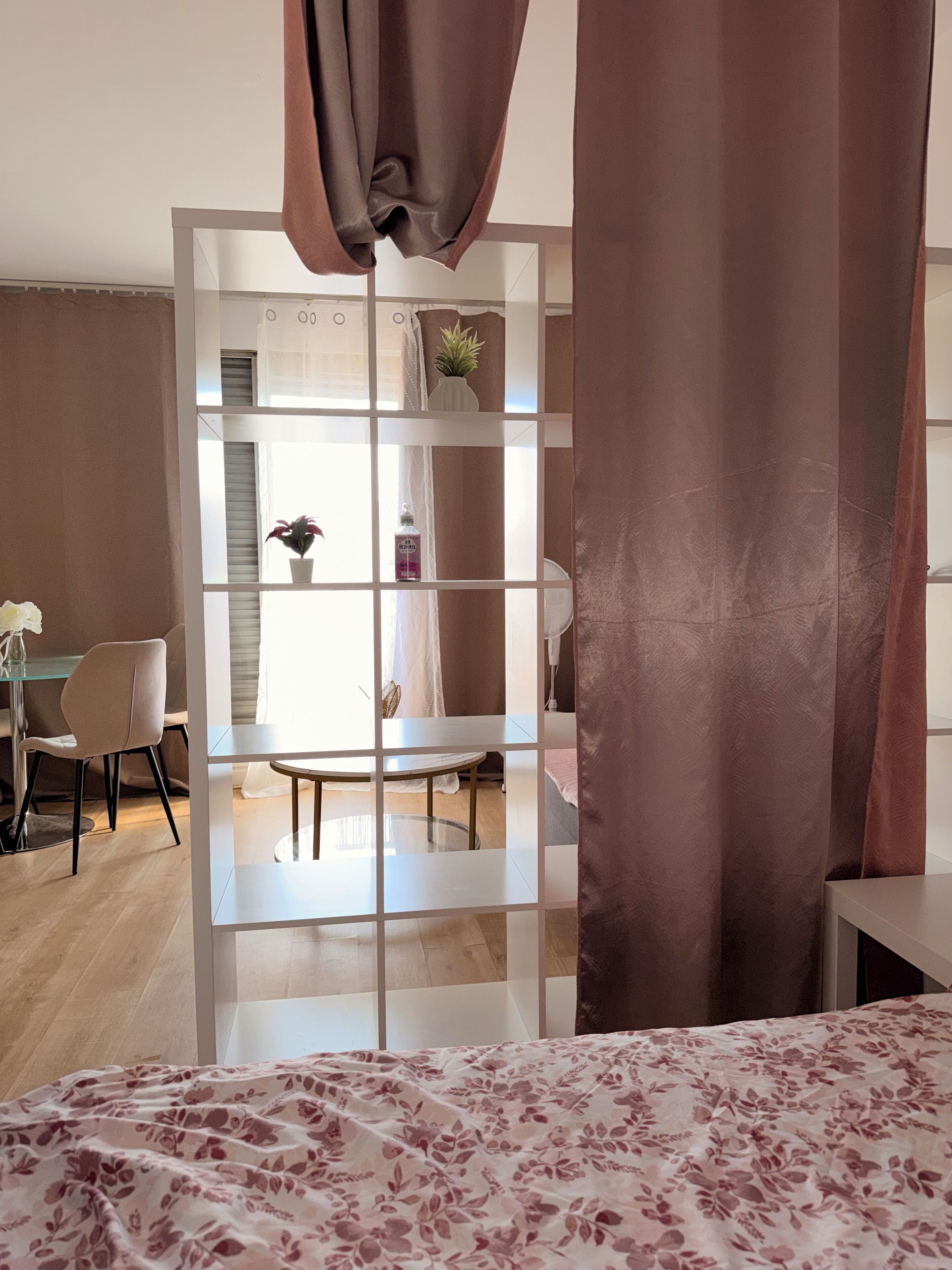 Habitación con cama, escritorio y silla. Cuenta con ventana con cortinas, una planta en el escritorio y una botella de loción.