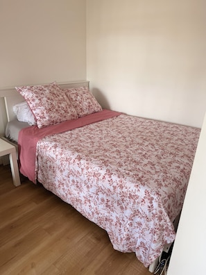 Uma cama de solteiro com colcha florida, uma mesinha de cabeceira e um criado-mudo.