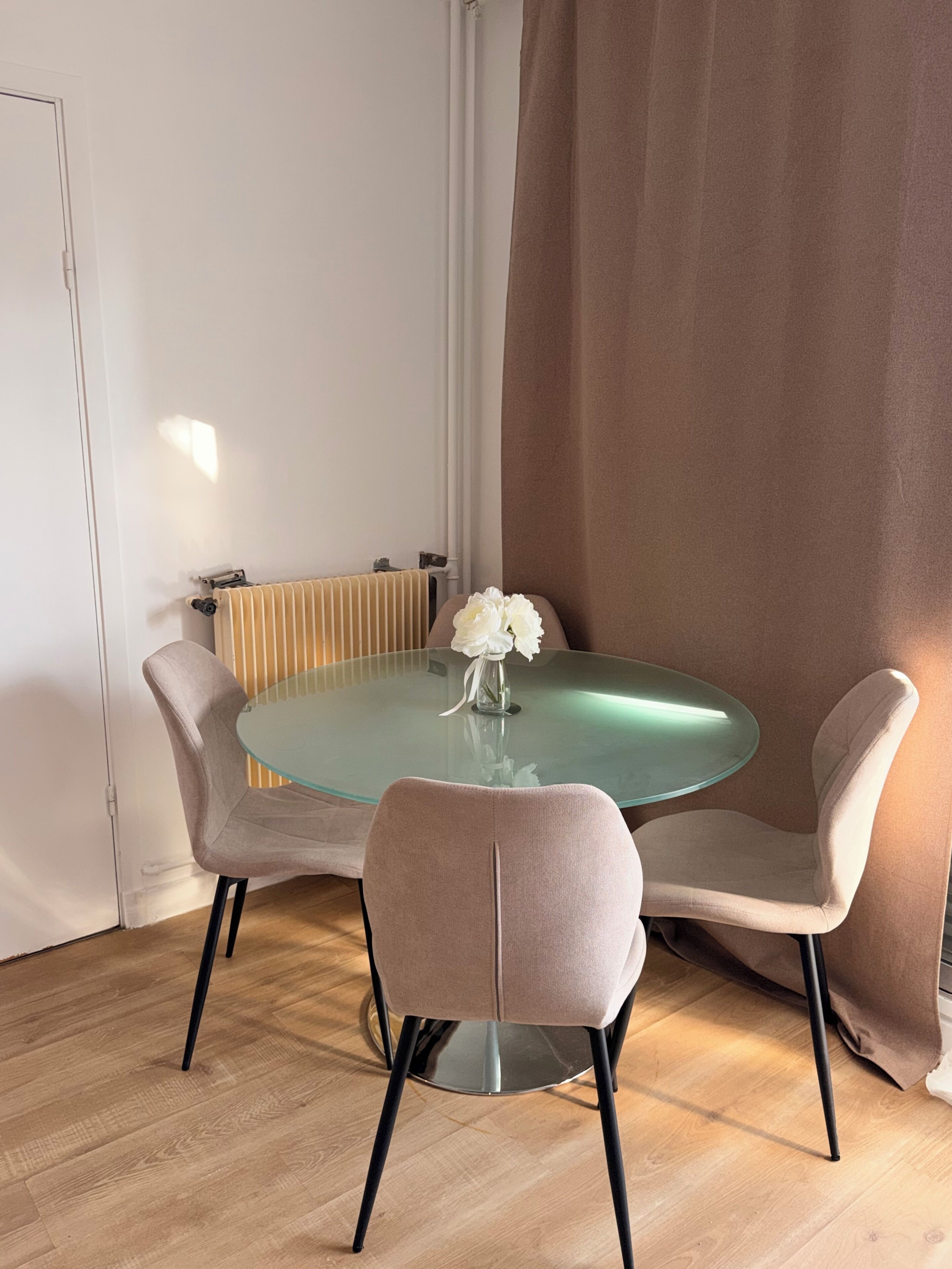 Un comedor con una mesa redonda de vidrio, dos sillas acolchadas y un jarrón con flores blancas.