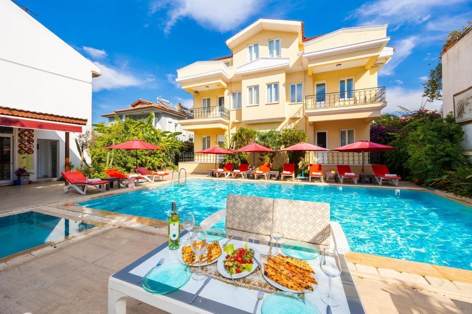 Zona de piscina con sillones de descanso y sombrillas, una mesa de comedor con platos y vasos, y un edificio con balcones.