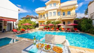 Zona de piscina con sillones de descanso y sombrillas, una mesa de comedor con platos y vasos, y un edificio con balcones.