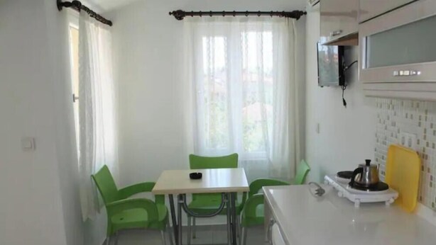 Una pequeña cocina con mostrador blanco, estufa y microondas. Hay una mesa con dos sillas y una ventana con cortinas.