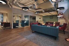 A spacious lounge area with a large sofa, multiple chairs, and a coffee table. There is a kitchen area in the background, and the ceiling features exposed ductwork and lighting fixtures. - Student Only Picturehouse Apartments (Exeter)