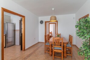 A dining area with a wooden table and chairs, a hanging light fixture, a clock on the wall, and a refrigerator in the adjacent kitchen.