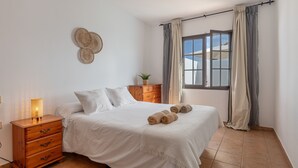 A bedroom with a bed, a nightstand, a lamp, a window with curtains, and a wall decoration. - Vacation Home 'Timanfaya' with Sea View, Private Pool and Wi-Fi (Tías)