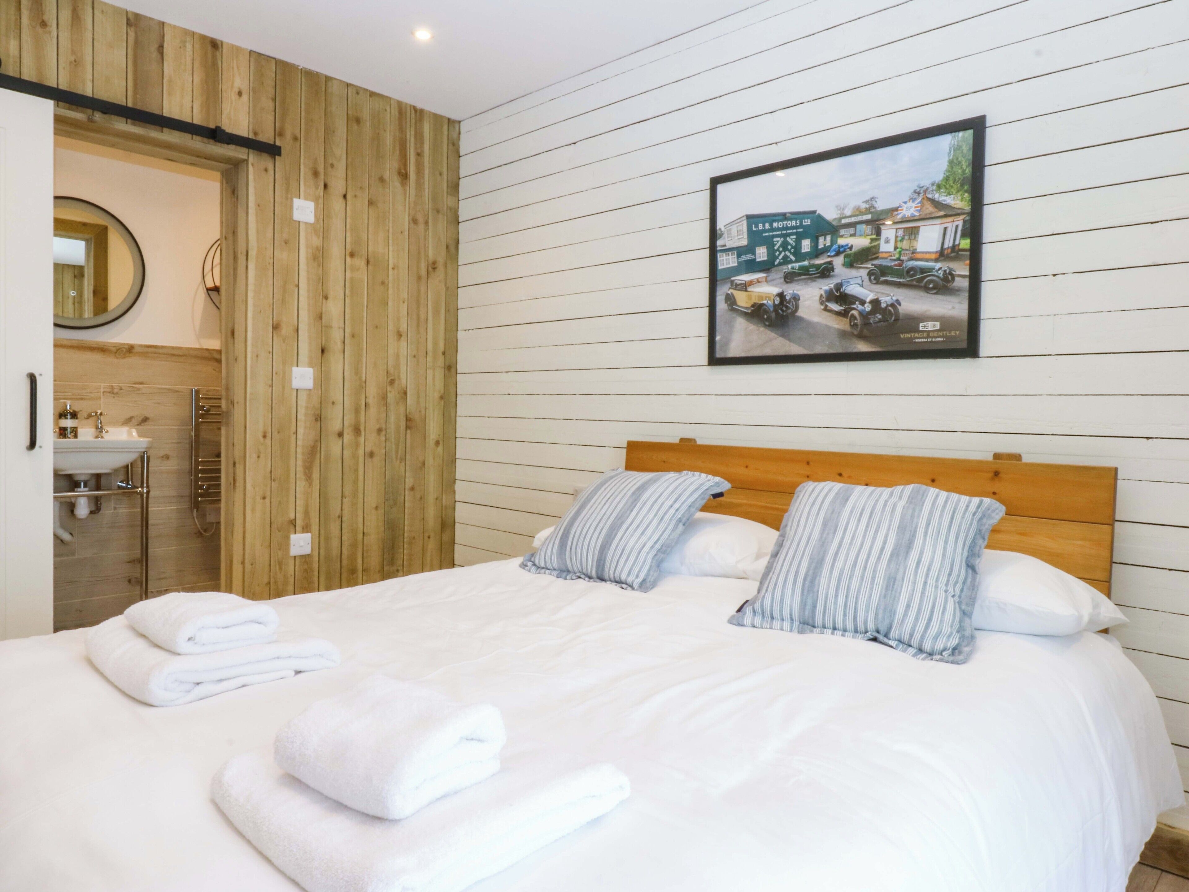 Una cama bien hecha con sábanas blancas y dos almohadas, un cabecero de madera, un cuadro en la pared y paneles de madera.