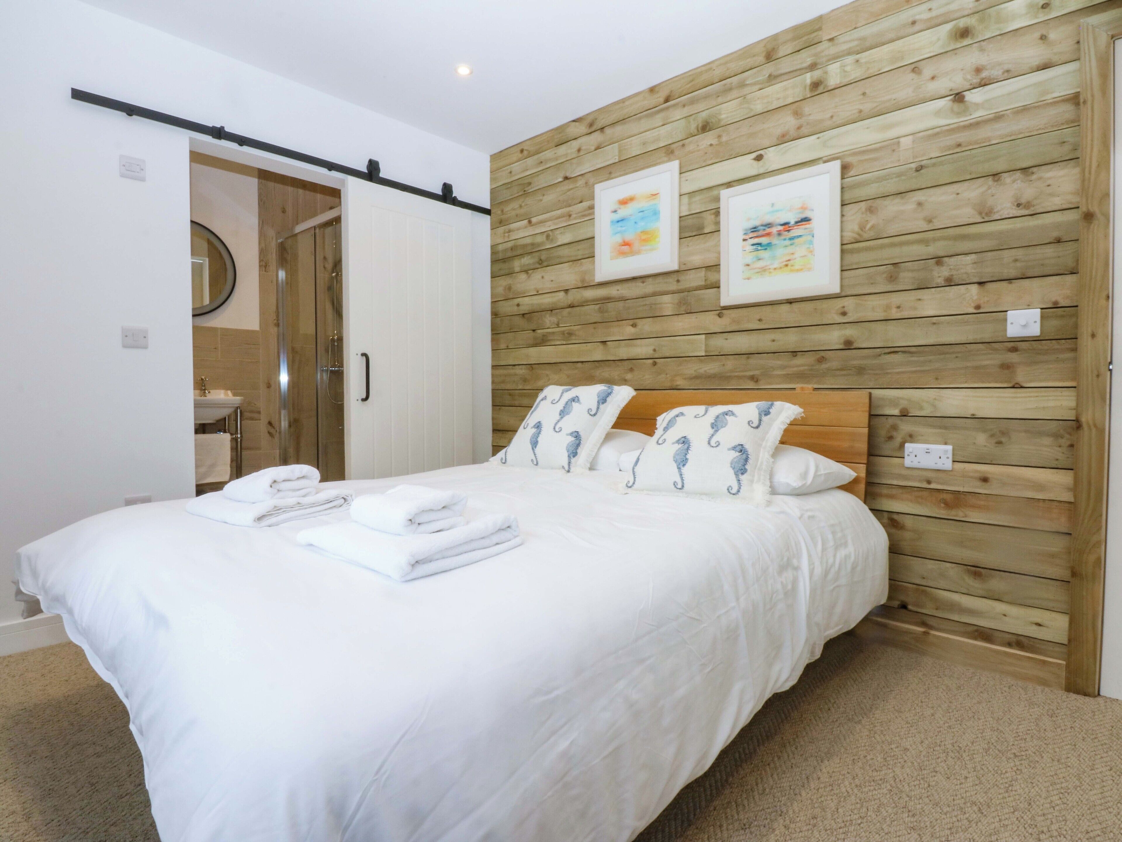 A bedroom with a wooden wall, a bed with white bedding and blue pillows, and a bathroom visible through an open door.