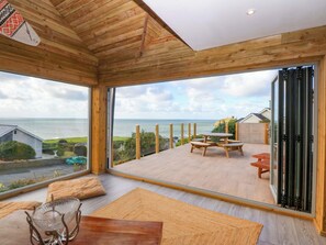 A modern interior with a large sliding glass door leading to an outdoor deck. Inside, there is a wooden table with chairs, a glass bowl on a stand, and a wooden bench. - Gowan Brae (Criccieth)