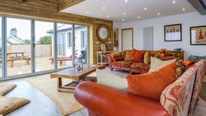 Un vaste salon doté d’une grande porte-fenêtre coulissante donnant sur une terrasse extérieure, d’un plafond en bois et d’un coin salon confortable avec un canapé et des coussins à motifs.
