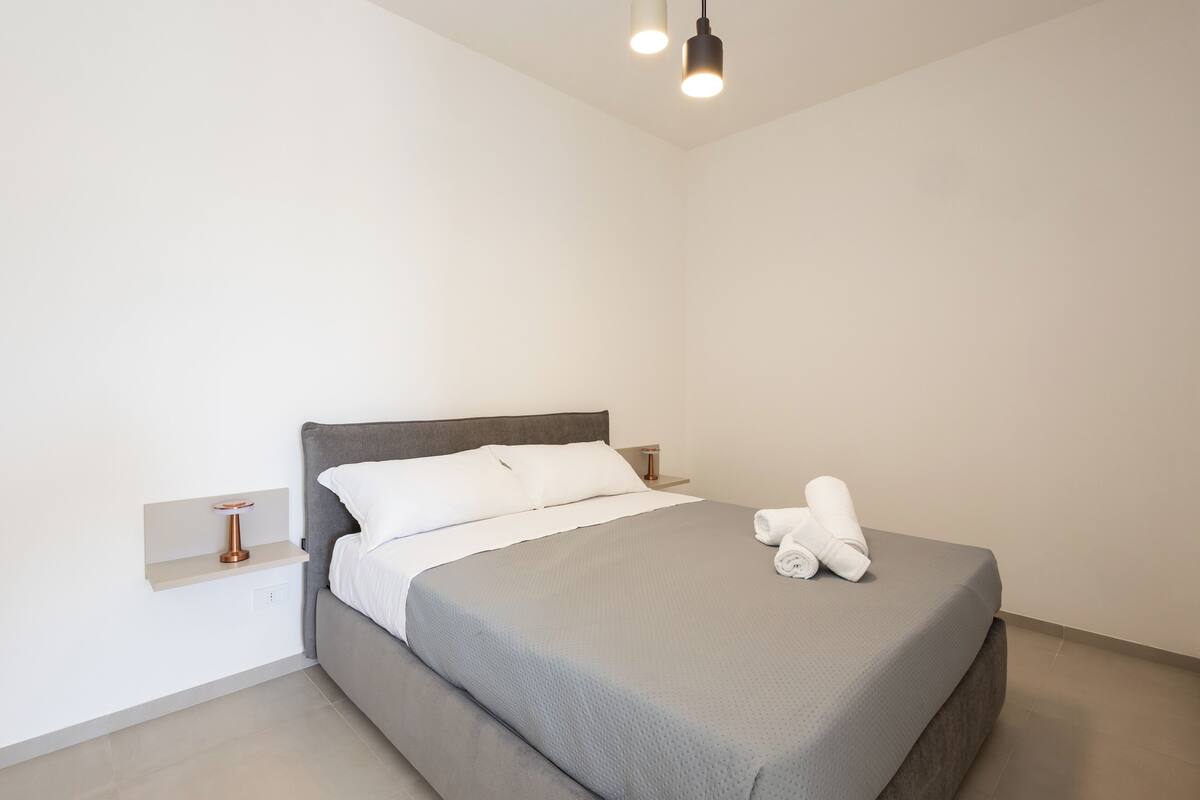 Ein modernes Schlafzimmer mit einem grauen Bett, weißen Kissen und einem ordentlich bezogenen Bett mit Handtüchern.