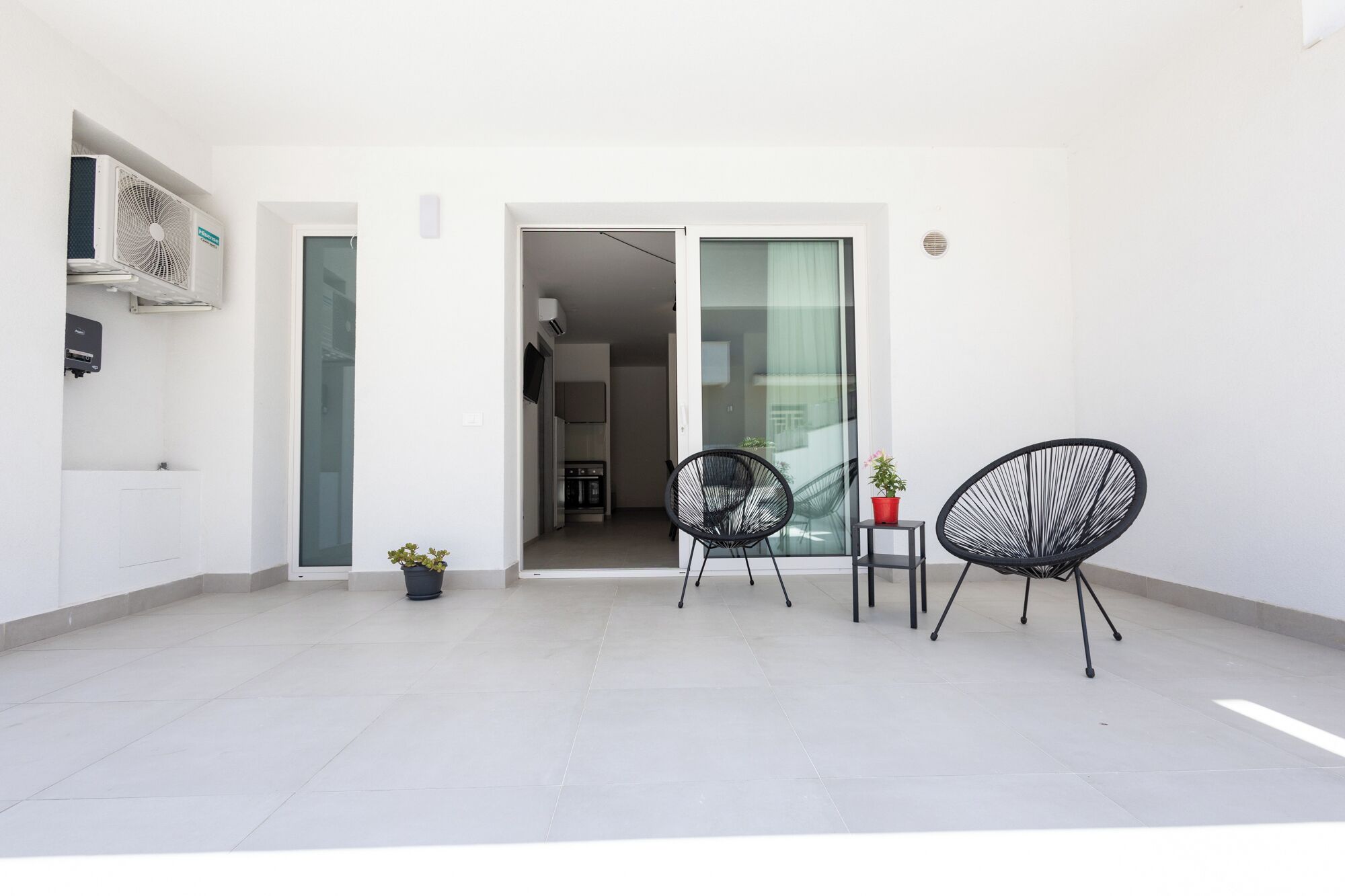A modern apartment with a balcony, two black wireframe chairs, a small table, and a potted plant.