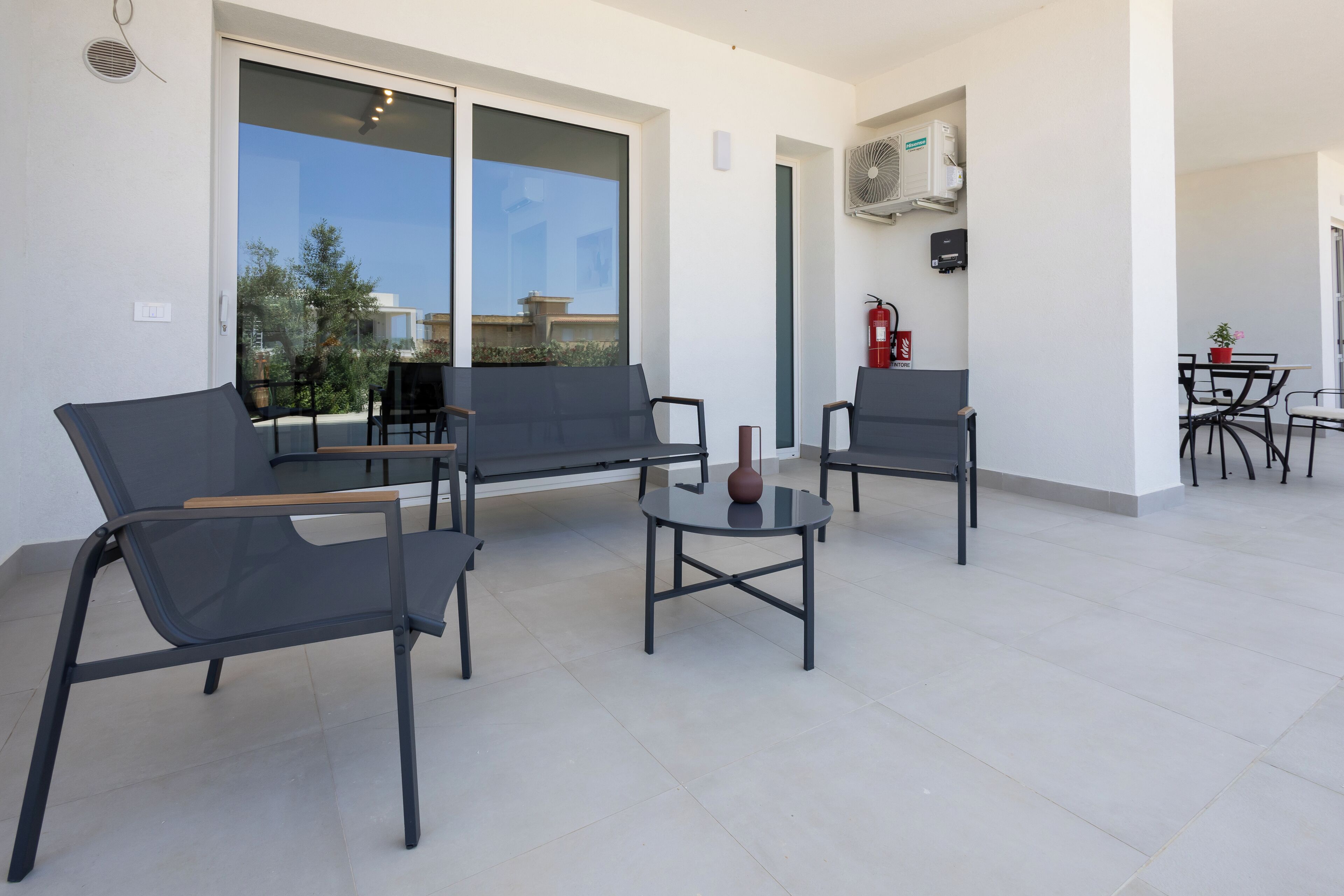 Een modern buiten zitgedeelte met zwarte stoelen en een kleine ronde tafel, grote ramen met uitzicht op de stad, en een airconditioning unit aan de muur.