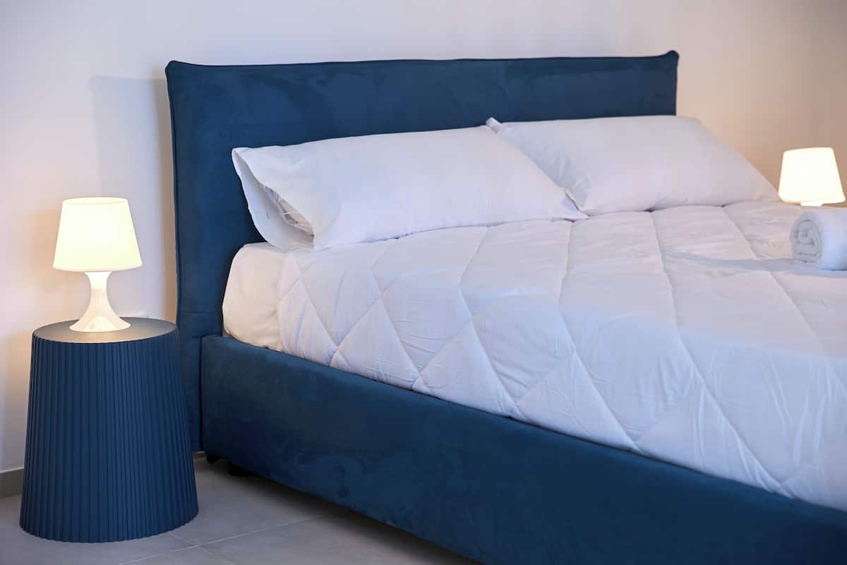 Ein ordentlich bezogenes Bett mit einer gesteppten weißen Bettdecke, zwei Kissen und einem passenden blauen Kopfteil. Auf dem runden Nachttisch steht eine Lampe.