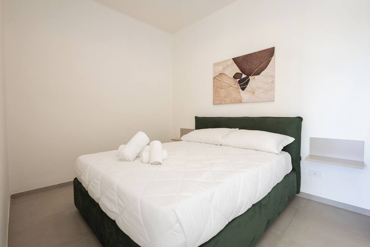 Ein ordentlich bezogenes Bett mit weißen Leinentüchern und einem grünen Kopfteil, ein an der Wand befestigtes abstraktes Gemälde und ein kleines Regal mit einem Objekt in hellem Farbton.