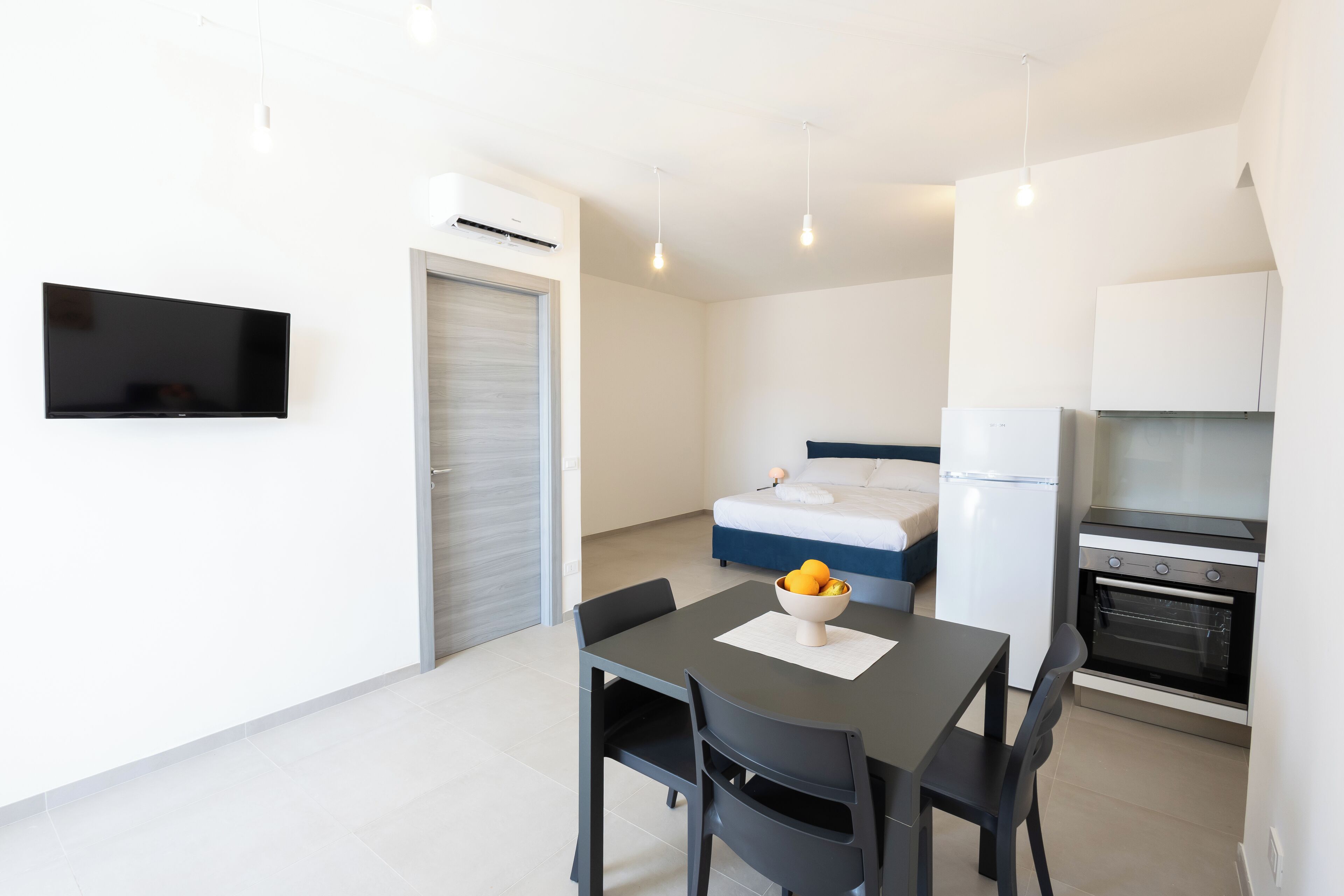 Une pièce moderne et bien éclairée, comprenant une table à manger, des chaises, un lit, un téléviseur et un coin cuisine.