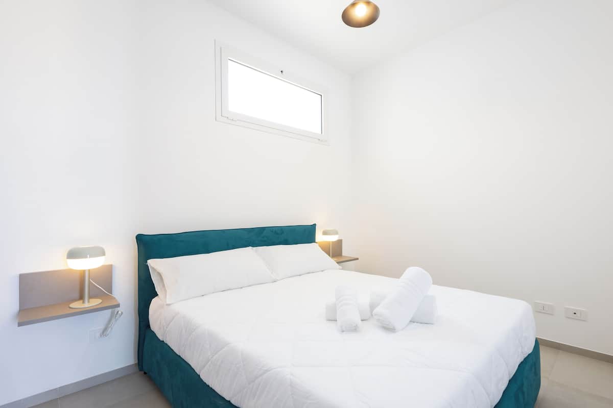 Ein ordentlich bezogenes Bett mit weißen Leinen und einem türkisblauen Kopfteil, flankiert von Nachttischen mit Lampen, in einem minimalistischen Raum mit großem Fenster und Deckenleuchte.