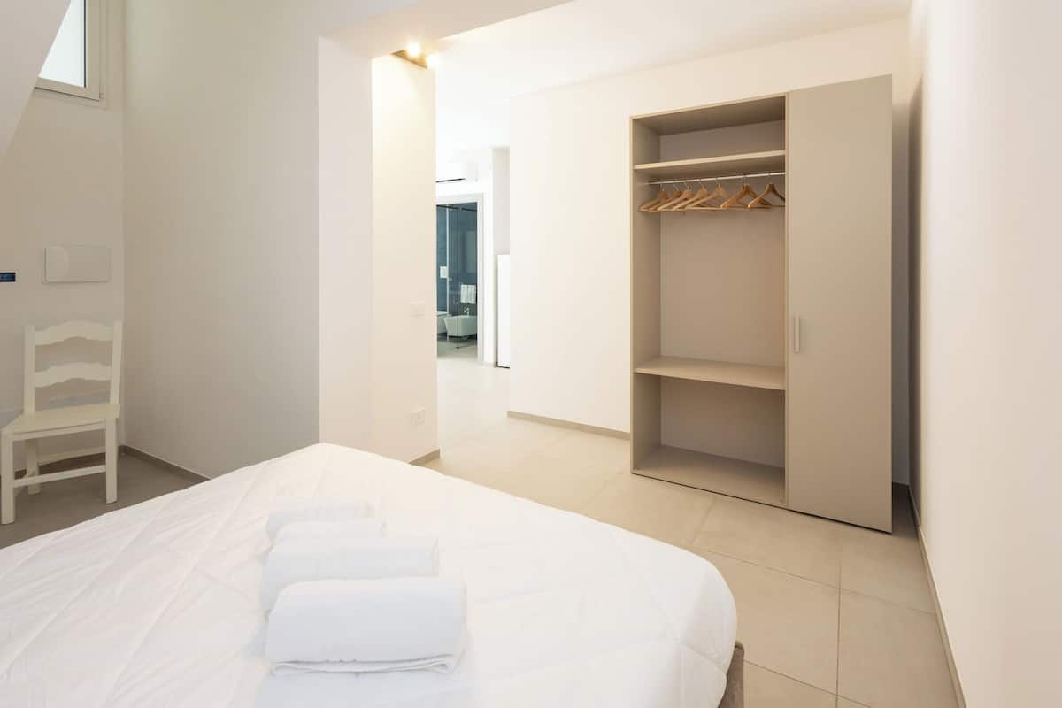 Ein Schlafzimmer mit Bett, einem Holzstuhl, einem Kleiderschrank und einem durchgehenden Türrahmen sichtbaren Badezimmer.