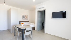 Una habitación de hotel moderna y minimalista con zona de comedor, cama con cabecera, televisor de pantalla plana en la pared y equipo de aire acondicionado.
