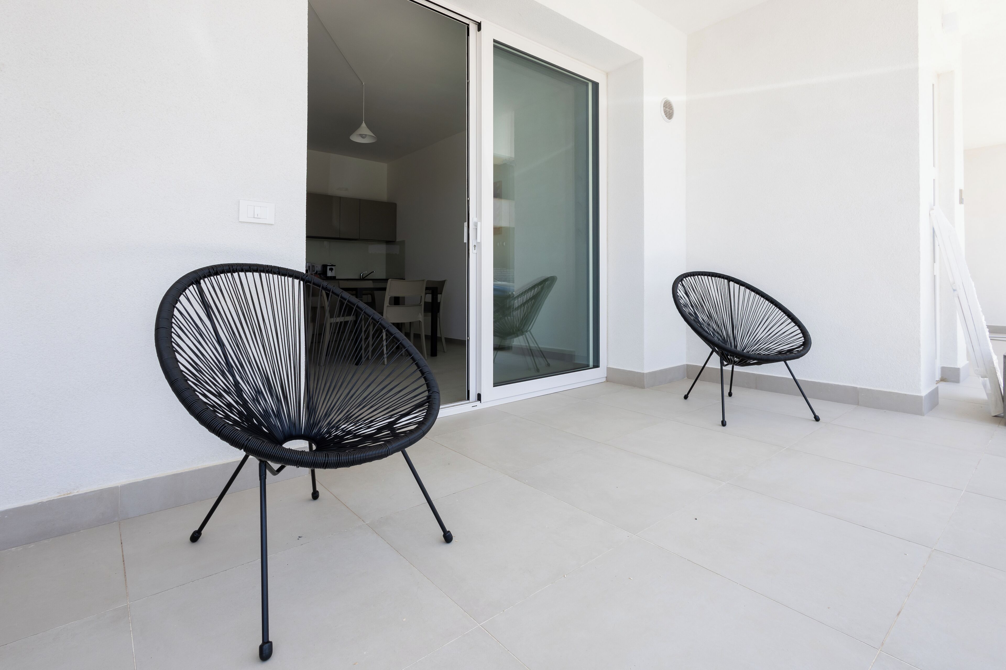 Un balcón moderno con dos sillas de estructura metálica negra y una puerta corredera de vidrio que da a un espacio interior.