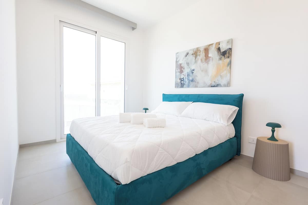 Ein modernes Schlafzimmer mit einem blaugrünen Bett, weißer Bettwäsche, einem kleinen Nachttisch und einem an der Wand befestigten abstrakten Gemälde.