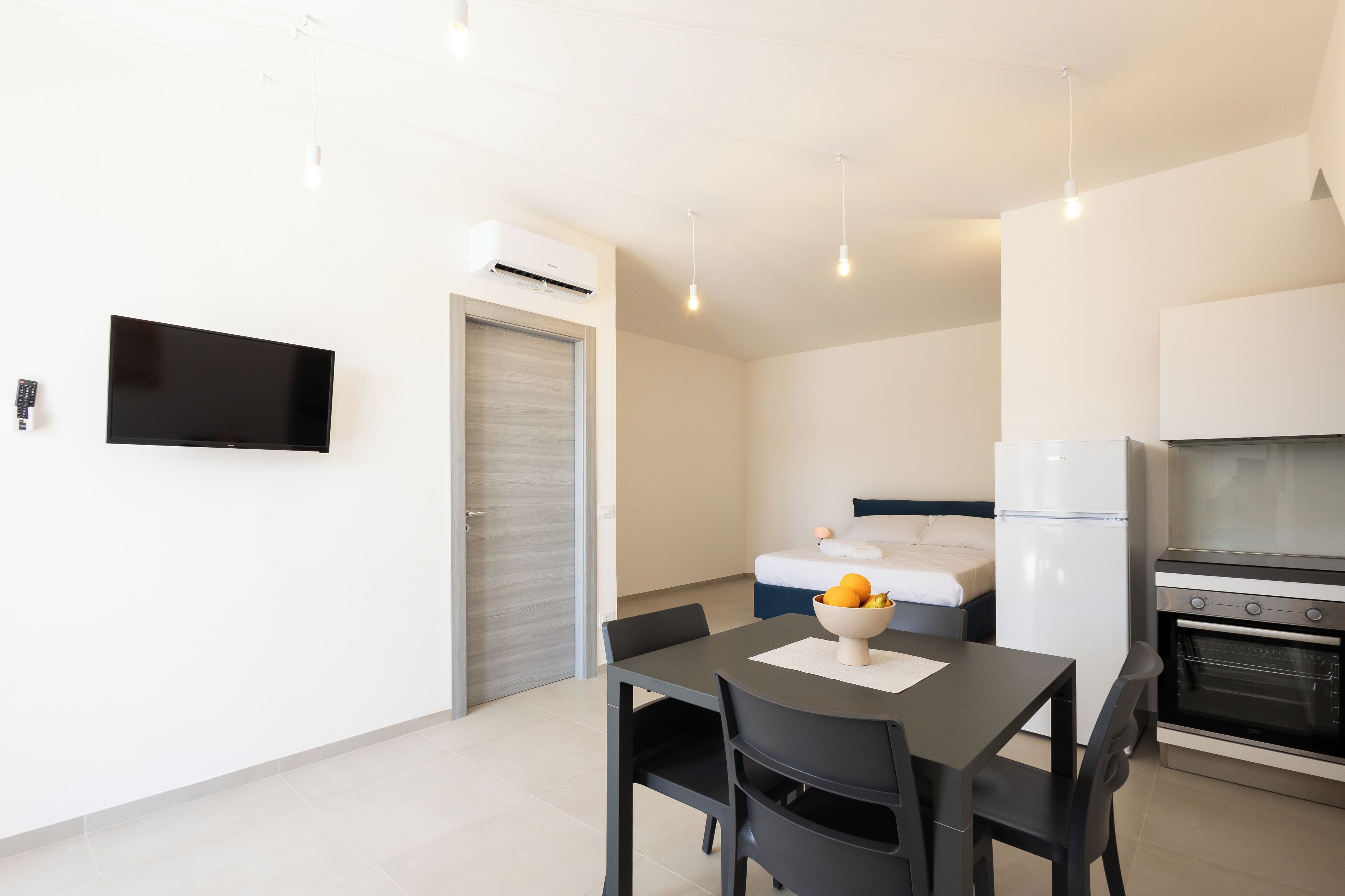 A modern, minimalistic room with a dining table, chairs, a TV, an air conditioning unit, a bed, a refrigerator, and an oven.