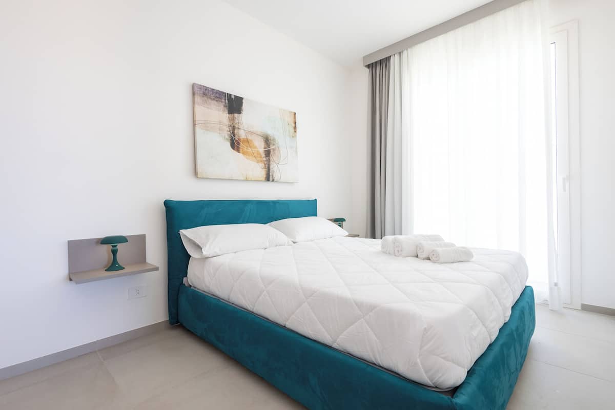 Ein modernes Schlafzimmer mit einem türkisfarbenen Bettgestell, weißer Bettwäsche, einem an der Wand befestigten abstrakten Gemälde und einem Fenster mit durchsichtigen Vorhängen.