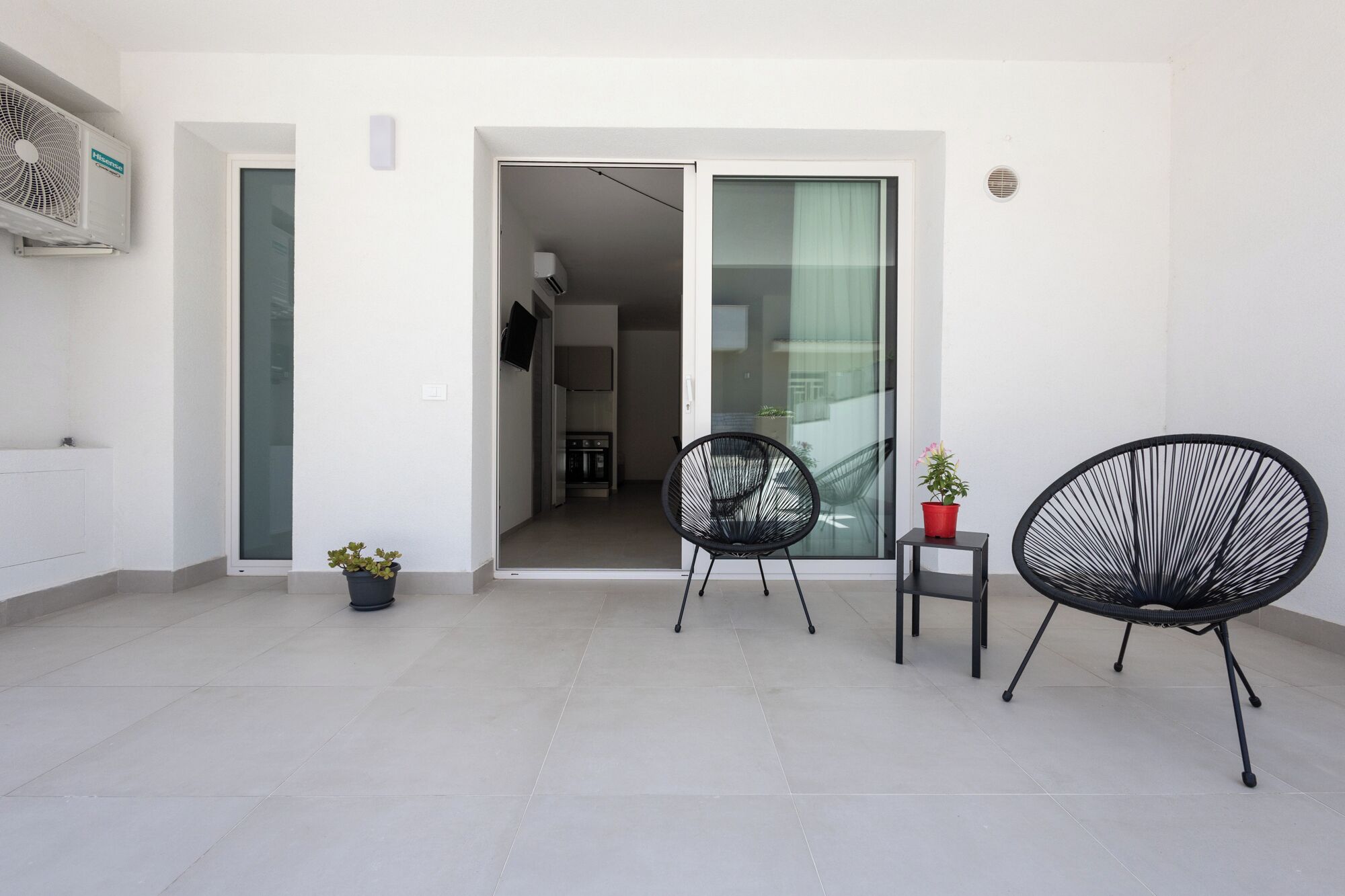 A modern, minimalist balcony with two black wireframe chairs, a small potted plant, and a sliding glass door leading to an interior space.