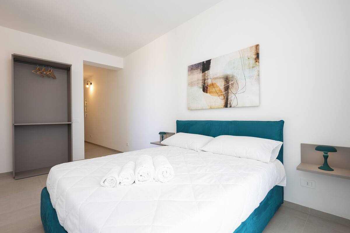 Ein modernes Hotelzimmer mit einem großen Bett, einer türkisblauen Kopfteil und einem an der Wand hängenden abstrakten Gemälde.
