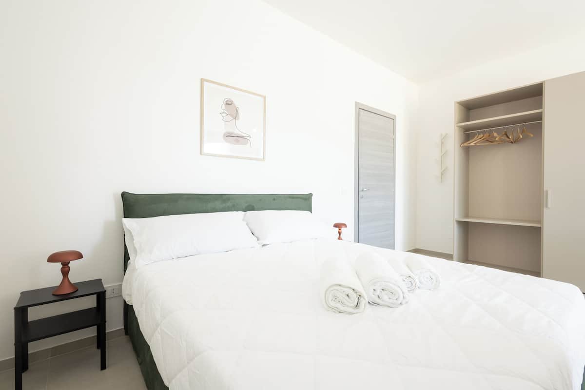 Ein ordentlich gemachtes Bett mit weißer Bettwäsche und zusammengerollten Handtüchern, ein Nachttisch mit Lampe, ein gerahmtes Bild an der Wand und ein Kleiderschrank mit Kleidung.