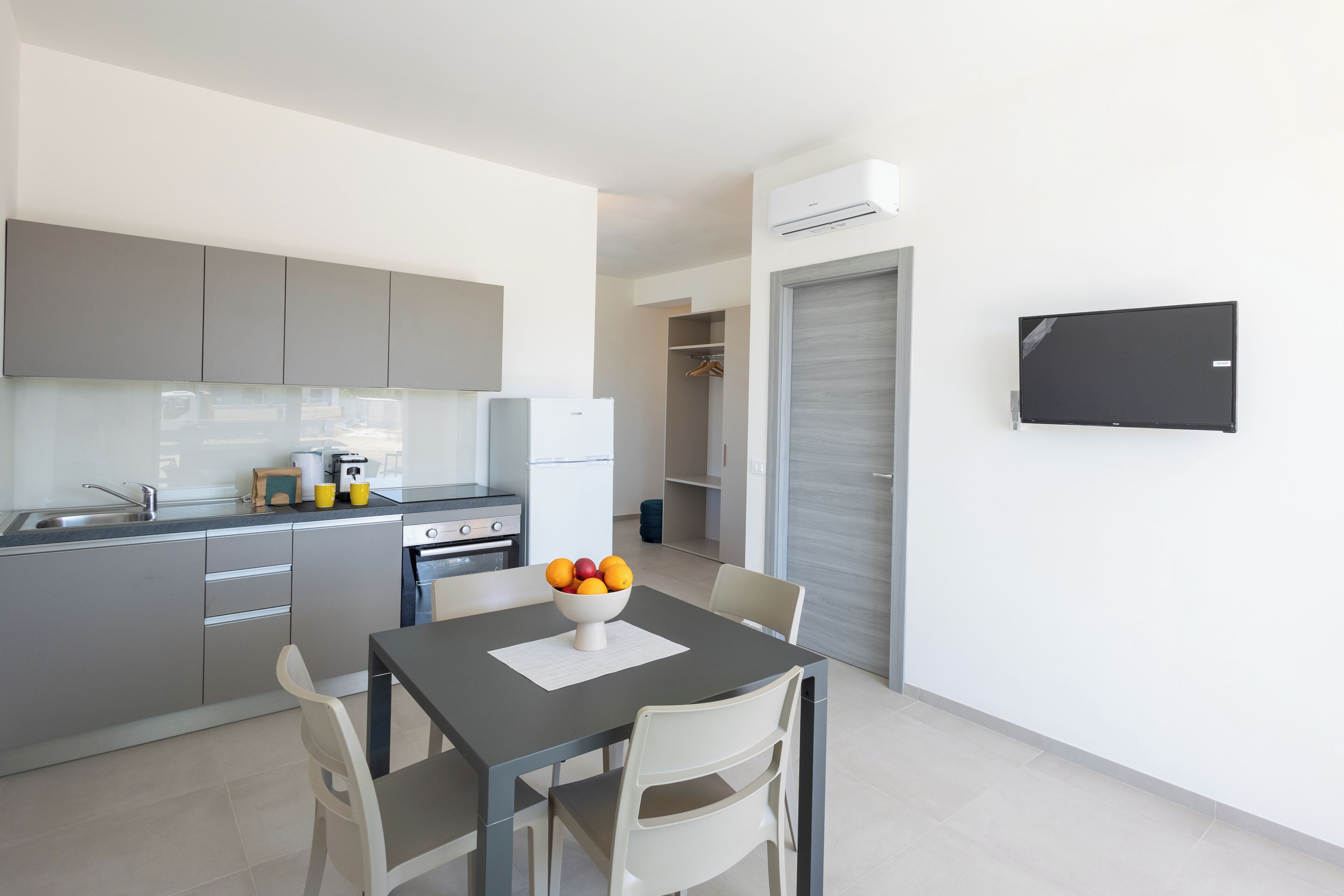 A modern kitchen with grey cabinets, a dining table with a bowl of fruit, and a TV mounted on the wall.