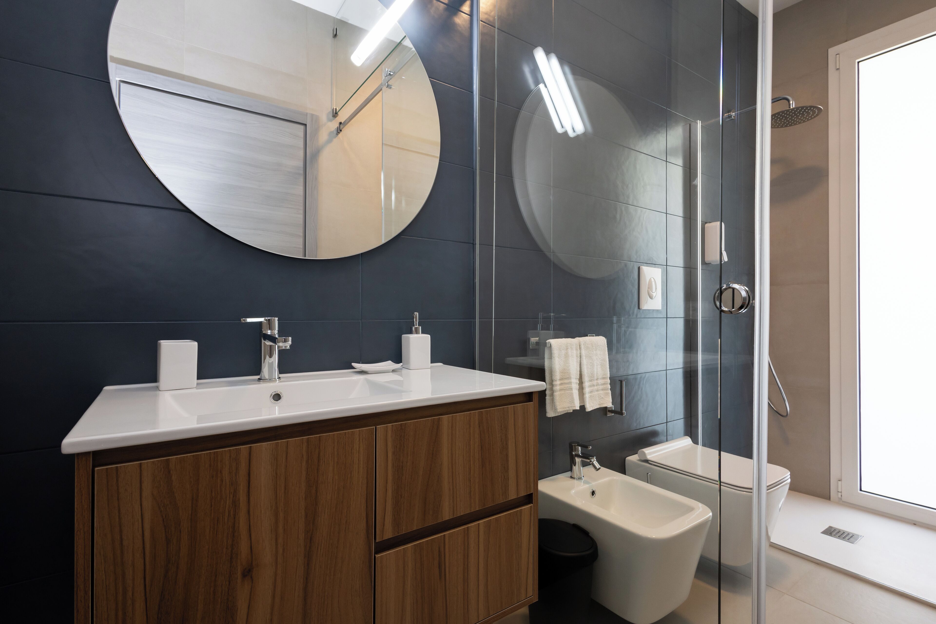 A modern bathroom with a round mirror, wooden vanity, white sink, and a glass-enclosed shower area.