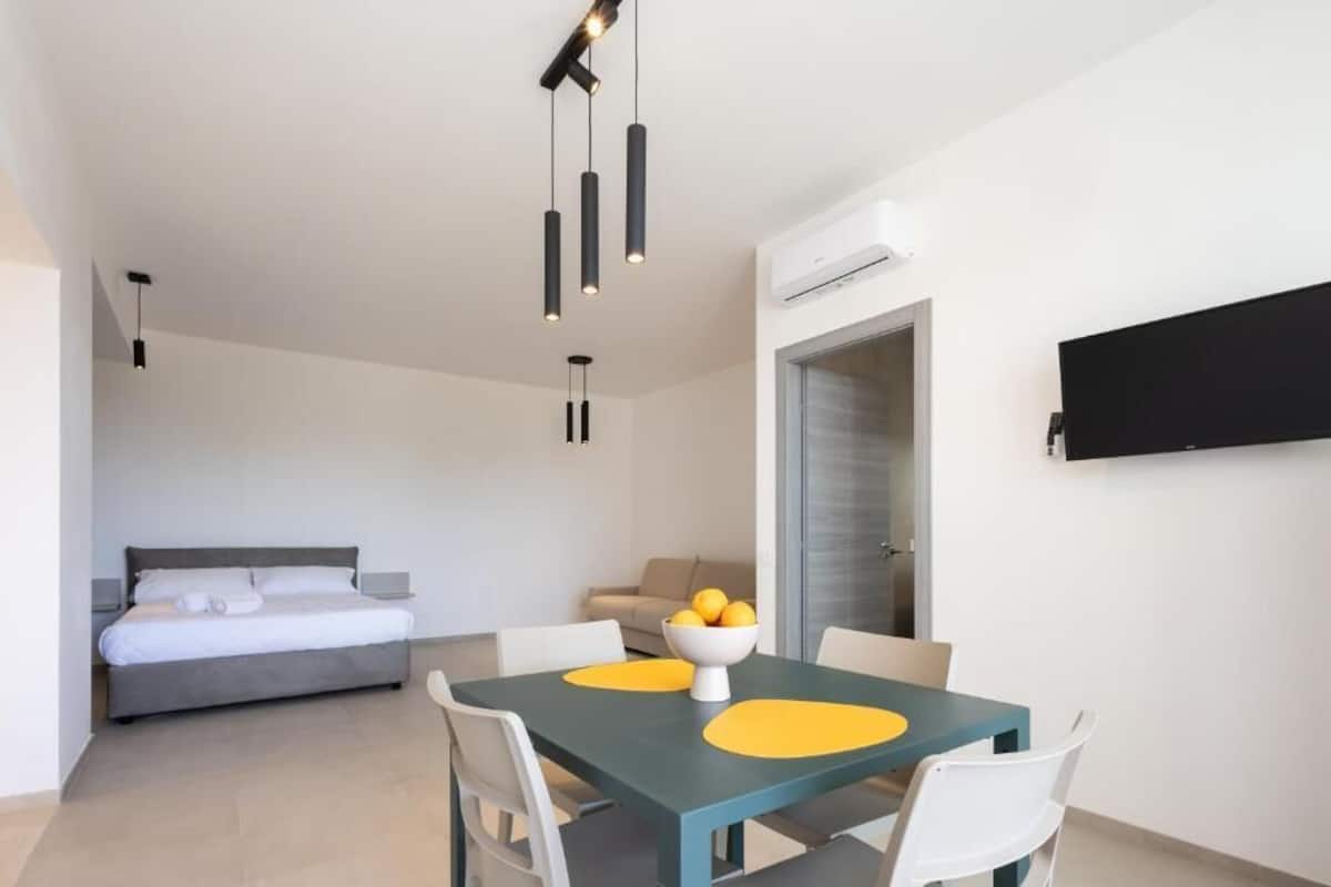 Ein modernes Hotelzimmer mit einem Bett, einem Esstisch mit gelben Platzsets, einer Obstschale und einem flachbildfernseher an der Wand.